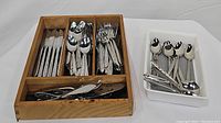 Wooden tray holding knives in one compartment, spoons in another, forks in a third, and serving utensils in the bottom compartment plus a white plastic tray with additional spoons and small forks.
