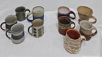 Overview photo showing all 10 ceramic mugs arranged on a white surface with diverse colors, patterns and textures visible.