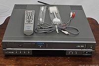 Front view of Samsung DVD-V2000 DVD VCR combo player with remote control and AV cables on top.
