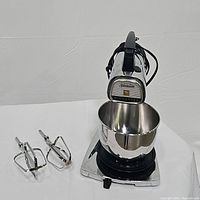 Front angled view of the vintage Sunbeam Mixmaster stand mixer with attached stainless steel mixing bowl and two stainless steel beaters placed beside the mixer on a white surface.