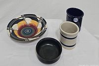 Four ceramic items: abstract multicolor serving bowl, black round serving bowl, tan crock with blue bands, and cobalt blue utensil crock displayed on white surface
