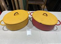 Two Great Jones cast iron pots side by side on a table, one yellow and one red, both with yellow lids with bronze handles and raised logos.