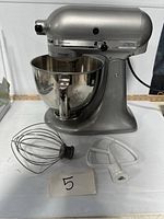 Photo showing the KitchenAid Artisan mixer with attached stainless steel bowl and two attachments (wire whisk and flat beater) placed in front.