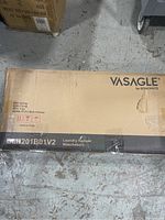 Photo of the closed original packaging box for Vasagle Songmics laundry hamper model BLH201B01V2 lying on a gray concrete floor, showing brand and model information.
