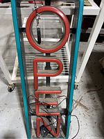 Frontal photo of rectangular illuminated open sign in teal frame with red letters.