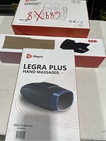 Photograph showing three LifePro product boxes stacked: one Legra Plus Hand Massager box, one Radial Leg Massager box, and one smaller additional box. The front face of the Legra Plus Hand Massager box is prominent with product image and branding.