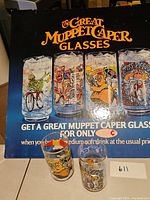 Photo showing promotional poster with title and images of four Muppet Caper glasses, and two collectible glasses placed in front.