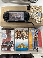 Sony PSP displayed with NBA Street Showdown on screen, charging cord, headphones, two game cases (FIFA Soccer, NBA Street Showdown), and two UMD movie cases (Super Troopers, Napoleon Dynamite) on cardboard surface.