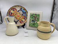 Lot overview showing two pitchers, colorful decorative platter on stand, and ceramic tile wall art mounted on white backing