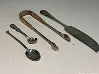 All four sterling silver utensils arranged side by side on white surface: two small spoons, tongs, and butter cutter.
