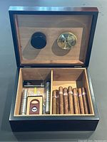 Humidor open showing interior compartments with cigar cutter, lighter, two carrying tubes, decorative cigars, hygrometer, and humidifier vent inside lid.