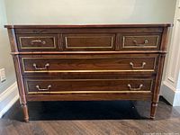 Front view of antique style walnut dresser showing three drawers with gold trim and brass handles on a dark wood floor against a light wall.