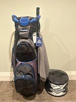 Front view of BagBoy golf cart bag with multiple zippered compartments and attached towel, next to black and white Impact Trainer carrying case.