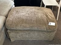 Side angle showing beige-tan chenille fabric ottoman with down filled cushion top and skirted sides on carpeted floor.