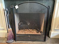 Full view of black metal fireplace screen in front of a fireplace with fire tools stand holding broom, poker, and shovel.