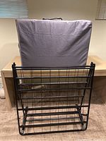 Two-piece folding single bed items, metal frame and folded mattress in carrying case, shown upright against a wooden table in a carpeted room.