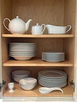 Shelf with stacked white porcelain plates, bowls, tea pot, sugar bowl, and gravy boat inside wooden cabinet.