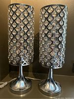 Pair of modern chrome table lamps with cylindrical shades covered in a metallic lattice housing plastic crystals, shown from the front against a dark wall.