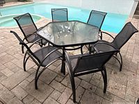 Full view of outdoor patio set with hexagonal glass table and six black mesh chairs arranged around it by a poolside on a paved floor.