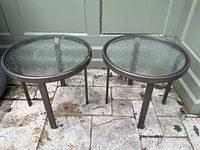 Photo showing two matching round outdoor side tables with glass tops and aluminum frames on a stone patio surface next to a wall.