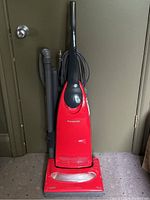 Front view of red Panasonic upright vacuum with attached hose and power cord bundled behind handle, showing full unit upright against a door.