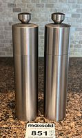 Front view of two stainless steel salt and pepper grinders side by side on countertop with auction sticker visible.