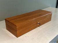 Closed solid wood rectangular jewelry box with polished warm wood tone finish and small gold lock with key inserted, placed on a white surface.