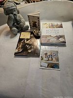 Concrete teddy bear sculpture seated on white fabric surface with several children's books and a small bell nearby
