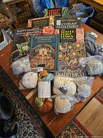 Wide view showing 7 needlepoint and embroidery design books and assorted yarn skeins spread on a wooden table.