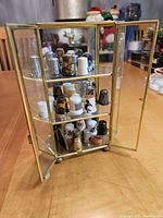Full view of the miniature curio display case with 23 assorted thimbles arranged on three glass shelves, showing a variety of colors and materials in bright lighting.