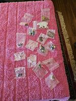 16 pairs of earrings packed in small plastic bags displayed on a pink textured fabric surface. Includes various designs such as a white polar bear, maple leaf, butterfly, floral, and pearl studs.