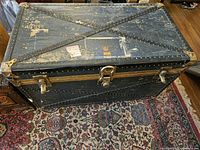 Top view of black trunk showing metal rivets and latches, surface has wear and paint loss.