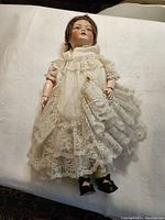 Full-length front view of doll on white background showing bisque head, lace dress, and black shoes.