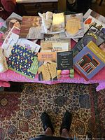 Photo showing 14 knitting books and 2 skeins of white yarn arranged on a pink surface with a patterned rug below.