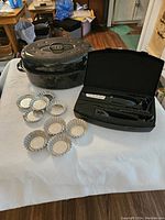 Photo showing all items in the lot displayed on a white cloth, including large black speckled enamel roast pan with lid, two black electric cutting utensils in case, and nine metallic tart forms.