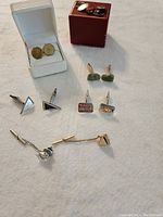 Image showing five sets of men's cuff links and two tie pins arranged on a surface with two boxes; white box contains hexagonal gold-tone cuff links, various colors and shapes visible