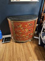 Full view of curved demi lune wooden dresser with floral painted decoration and brass hardware, placed on wood floor with dark wall behind.