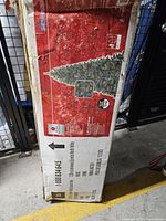 View of original cardboard box showing the artificial Christmas tree with some product information and branding indicating a 7.5 ft tall pre-lit artificial tree.