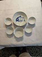 Top-down image showing one larger bowl with a fish motif and six smaller bowls arranged around it on white fabric.