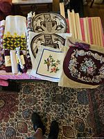 Wide view of assortment showing stretcher bars, embroidery canvases with owl designs, fabric pieces with floral needlework, and thread supplies on table.