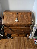 Front view of antique wooden writing desk with fold-down top closed, showing wood grain and decorative tassel on key lock.