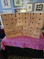 Five birch wood IKEA storage boxes arranged on a table covered with a purple cloth. Three boxes are square shaped, and two are wider rectangular shaped. Each box features multiple small drawers with finger cutouts.