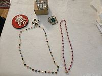 Six pieces of costume jewelry displayed on a beige cloth: three brooches, two necklaces, and one silver-tone cuff bracelet watch on box.