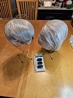Two gray styled wigs on metal stands with a pack of three black hair clips on a wood table.