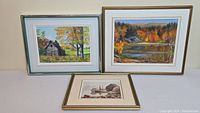 Three framed paintings displayed on wall: sailboat scene (bottom), cabin landscape (left), lake scene (right)