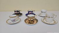 Six demitasse cups and saucers arranged in two rows on beige background, showing variety of colors and patterns