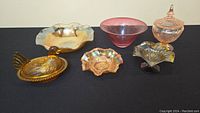 Full lot view showing six decorative glass pieces including cranberry glass bowl, carnival glass bowls, pink depression glass lidded jar, and amber glass hen dish.