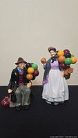 Two Royal Doulton porcelain figurines, a seated old man and a standing old woman, both holding bunches of colorful balloons