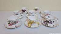 Full view of the 7 tea cup and saucer sets arranged in two rows, each set showing distinct floral patterns and gilded rims.