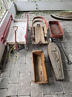 Overview photo showing all main items: large red metal wagon, wooden sled, small red wooden wagon, carved doll coffin, and wooden accessory/base piece.